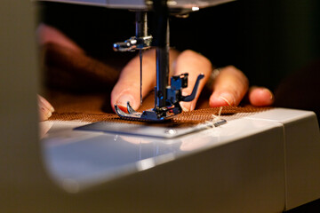 Mano cosiendo una tela marrón  con una maquina de coser iluminada por la luz de la propia máquina
