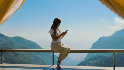 Woman holding a smartphone and scrolling through screen, sitting on a wooden terrace, looking out at the stunning mountain landscape. Female traveler is bathed in warm sunlight of a summer sunny day