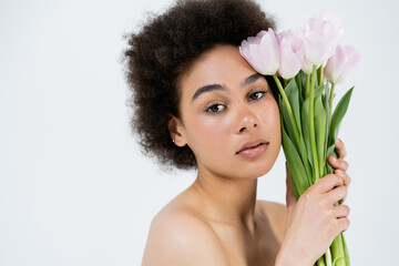 Portrait of african american model with naked shoulders holding tulips isolated on grey.