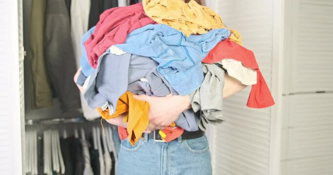 Woman with many clothes in hands turns to camera. Close-up, front view. Concept of unwashed linen, excess clothing, housework, overconsumption of things.