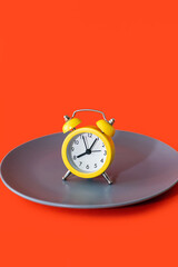 A yellow alarm clock stands on a plate on a orange vertical background. The concept of diet and proper nutrition on a schedule. Lunch break or meal time. Eating strictly at the agreed time