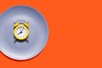 A yellow alarm clock lies on a plate on an orange background. The concept of following a diet and...