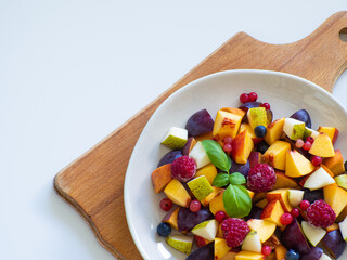 Delicious fruit salad with fresh plum, peach, nectarine, pear and frozen raspberries, decorated with green basil on white background. Top view. Healthy eating. Copy space. Vegan cuisine. Dietary menu.