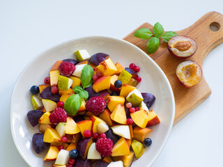 Delicious fruit salad with fresh plum, peach, nectarine, pear and frozen raspberries, decorated with green basil on white background. Top view. Healthy eating. Copy space. Vegan cuisine. Dietary menu.