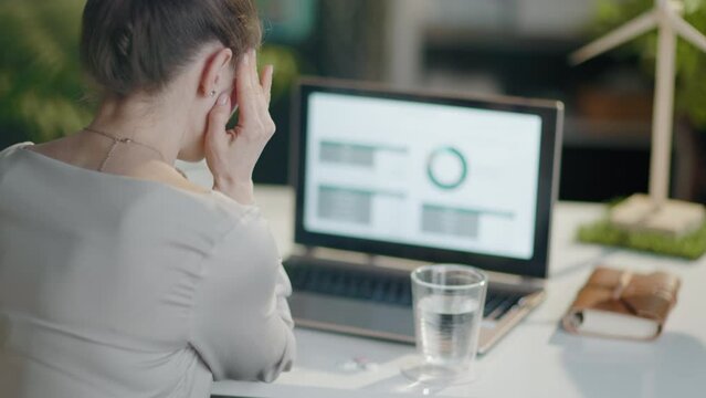 Seen from behind modern business woman with laptop, pills and glass of water having headache in modern office.