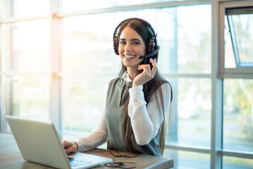 Portrait of friendly female customer support representative in headset with microphone consulting client, using laptop and looking at camera in customer support service office.