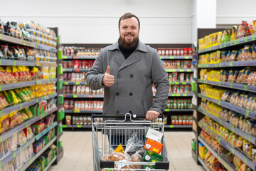 Smiling, contented young man with a beard and a gray coat stands in a grocery supermarket with a shopping cart filled with food and gives a thumbs up