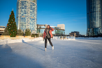 a young beautiful girl on skates in a sheepskin coat on a skating rink against the backdrop of the...