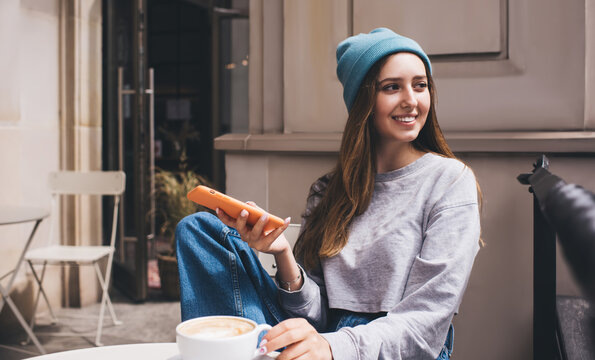 Young millennial female using application on mobile phone while drink coffee. Caucasian hipster girl holding smartphone in stylish silicone case. Teenager with cellphone enjoying free time in city