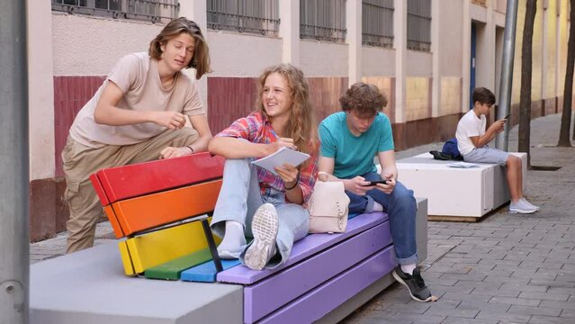 Teenager boy with smartphone and girl with copybook sitting on rainbow-colored bench. Their friend standing beside and talking with them.