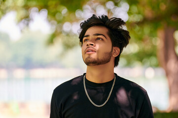 Attractive young indian man portrait in black t shirt and silver neck chain on outdoor green park...