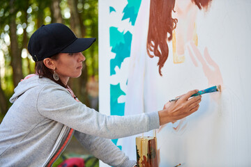 Girl artist draws with paint brush surreal woman portrait on white canvas at outdoor art painting festival, paintings art picture process. Woman in black cap paints atmospheric surreal picture - obrazy, fototapety, plakaty