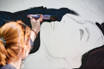 Woman artist draws with paint brush surreal girl portrait on white canvas at outdoor art painting festival, paintings art picture process. Woman in black cap paints atmospheric surreal picture - obrazy, fototapety, plakaty