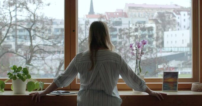 Woman stands at the window with her back and looks into the distance. Dullness and cold outside and warm inside. An unrecognizable person thinks about his destiny. Female existential midlife crisis.