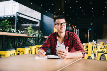 Cheerful freelancer with smartphone and notepad looking away