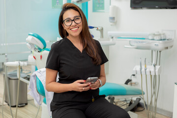Young female dentist looking at camera smiling, sitting in her office with a cell phone in her hand
