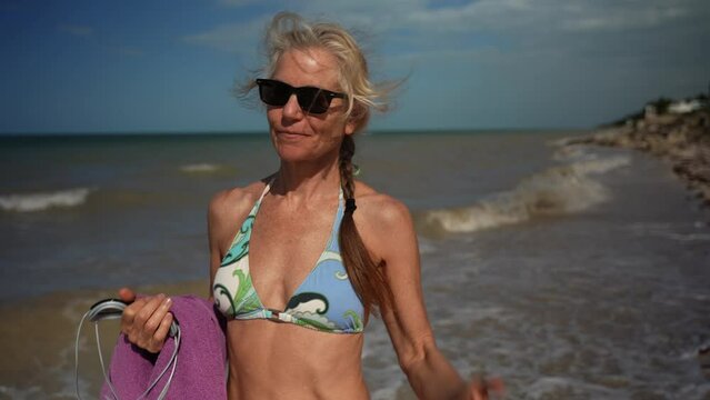 Closeup front view of happy smiling mature senior woman 50s, 60s, in bikini and sunglasses, walking on windy beach carrying towel and swim goggles with crashing waves behind her.