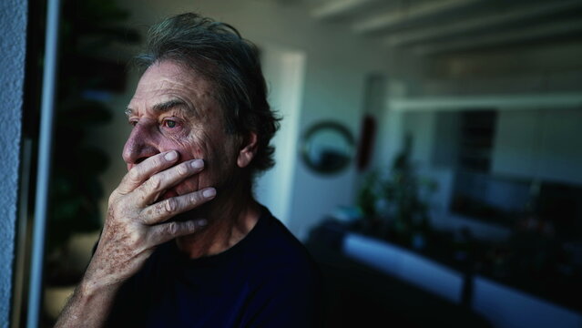 A fearful senior man standing by window with contemplative expression. Anxious expression of a mature older man in 70s at home indoors. apprehensive emotion