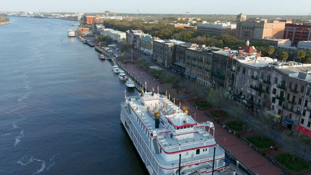 aerial footage flying along River Street with shops, restaurants, people walking, boats, ships and yachts docked along the banks and lush treen trees of the Savannah River in Savannah Georgia USA