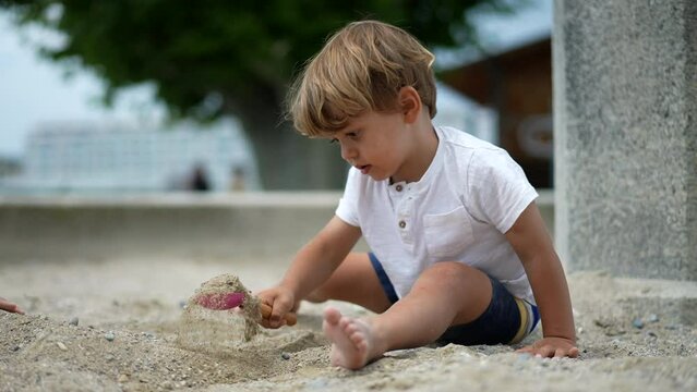 One little boy sitting outside at sandbox playing with shovel and sand. Child playing outdoors in summer day