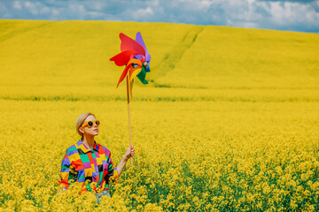 Beautiful female in 90s stylish shirt with pinwheel in rapeseed field