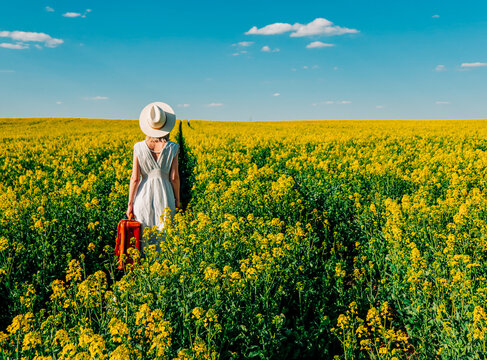Beautiful woman in dress with suitcase in rapeseed field in spring time