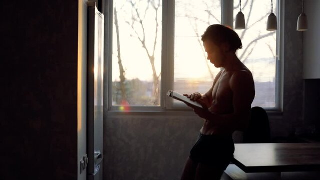man with a naked torso reads a book at home by the window.