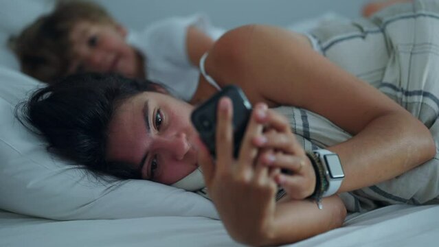 Woman lying in bed looking at phone in the morning. Candid lifestyle mother checking cellphone screen. People addicted to technology