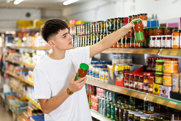 Focused thoughtful young guy choosing food in supermarket, holding glass jars with canned goods and...