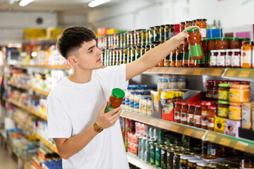 Focused thoughtful young guy choosing food in supermarket, holding glass jars with canned goods and reading labels