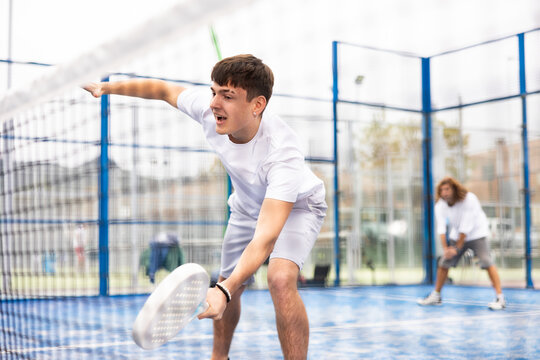 Portrait of emotional young sporty guy playing paddle tennis with friends on open court. View through net