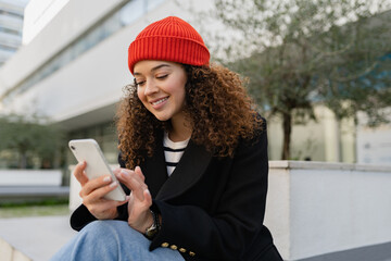 pretty curly woman in city street in autumn coat, using smart phone, red knitted hat