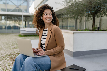 pretty curly woman sitting in city street in stylish jacket, working remote job on laptop