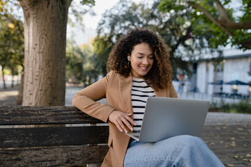 pretty curly woman sitting in city street in stylish jacket, working remote job on laptop