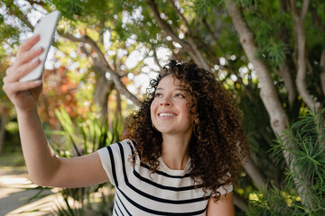 pretty curly woman walking in city street in striped t-shirt, using smart phone