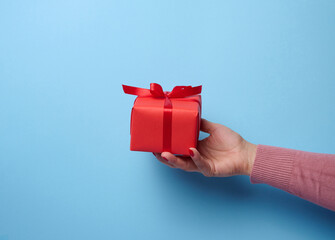 A woman's hand holding a red gift box wrapped with ribbon on a blue background, a concept of congratulations, surprise