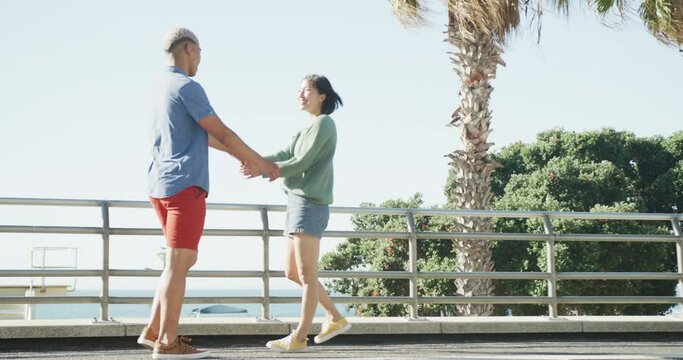 Happy biracial couple walking together and holding hands on promenade, in slow motion
