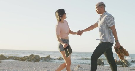 Happy biracial couple walking and holding hands at beach, in slow motion