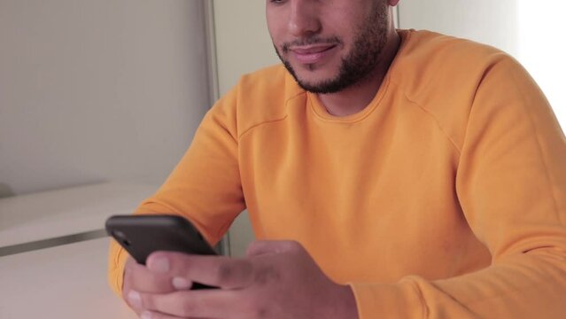Latin man sitting texting on cell phone smiling. Using cell phone in video call.