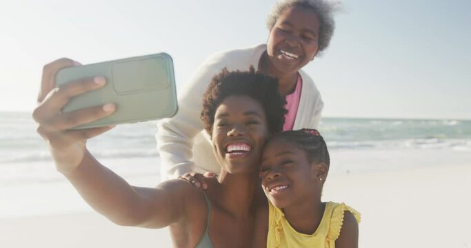 Happy african american grandmother, mother and daughter taking selfie at beach, in slow motion