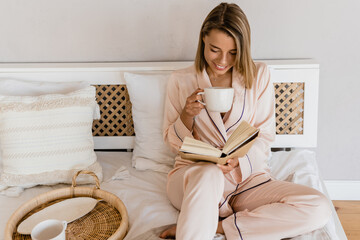 pretty smiling woman relaxing at home on bed in morning in pajamas reading book - obrazy, fototapety, plakaty
