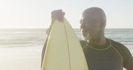 Happy senior african american man holding surfboard at beach, in slow motion