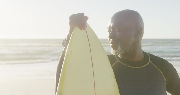 Happy senior african american man holding surfboard at beach, in slow motion