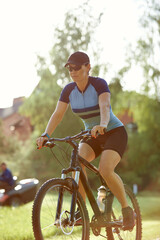 Smiling middle aged woman in cap with glasses cycling in summer park, cycling cardio training, city...
