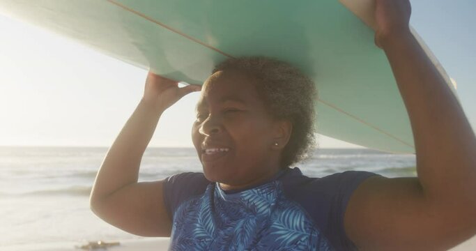 Happy senior african american woman walking and holding surfboard at beach, in slow motion