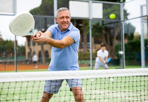 Sporty mature man padel player hitting ball with a racket on a hard court