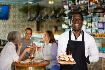 Happy African American waiter standing with plates of pinchos in the middle of the bar