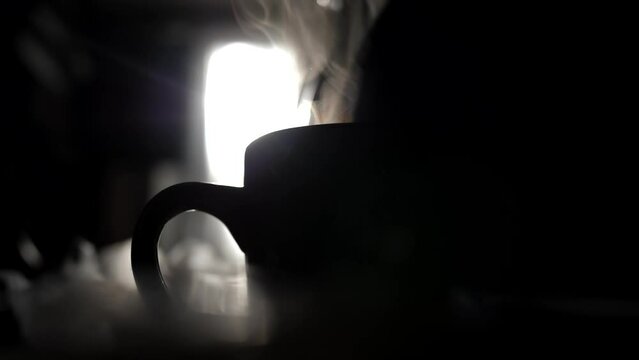 The silhouette of a cup of tea in the backlight, into which boiled hot water is poured from the kettle. Steam rises over a cup of hot drink during breakfast in the early morning.
