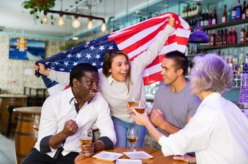 Cheerful american multiracial male and female celebrating spots team victory, waving flag of United States of America in beer bar