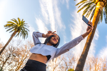 Young african woman posing in white clothes in a tropical place with palm trees at sunset taking a selfie